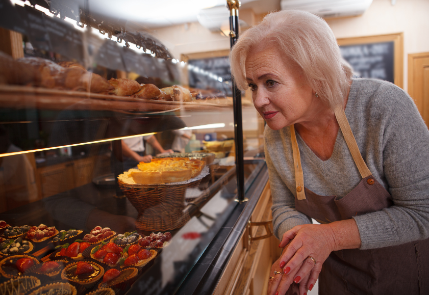 Older employee working in a bakery, smiling as she gets ready to take a pastry from display