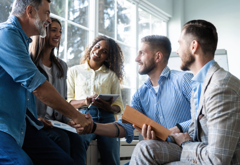 An employer shaking hands with employee amongst a smiling group of other office workers